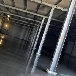 Gładka, szara powierzchnia w hali przemysłowej z metalowymi słupami i konstrukcją dachu, widoczne odbicia światła w posadzce.