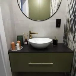 Nowoczesna łazienka z okrągłym lustrem, białą umywalką nablatową, złotą armaturą i szafką w kolorze oliwkowym, uzupełniona dekoracjami w postaci świec i dyfuzora zapachowego.