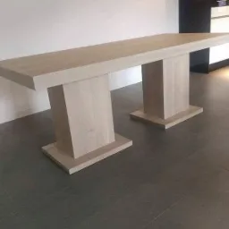 Stół z jasnego drewna o minimalistycznym designie, wsparty na dwóch masywnych, prostopadłościennych nogach z podstawami, na tle szarej posadzki i jasnej ściany.