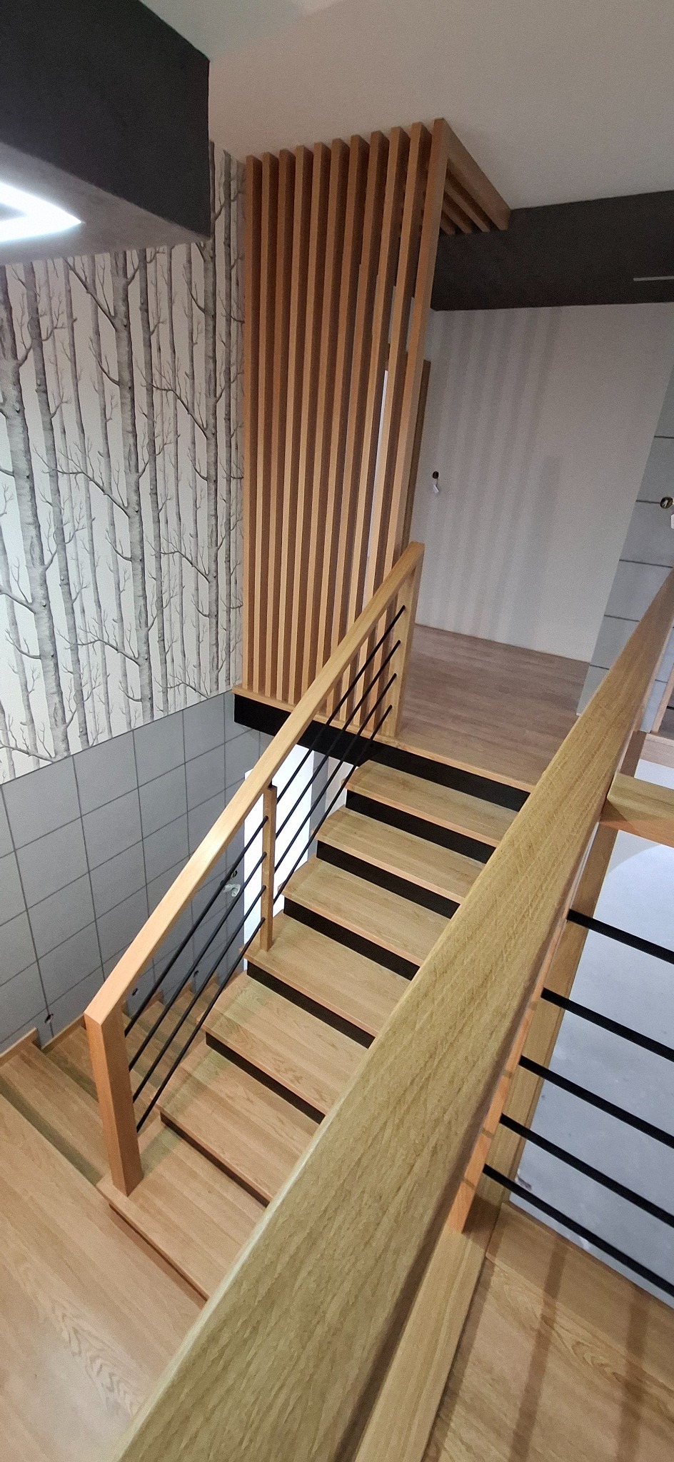 Nowoczesne schody o jasnym odcieniu drewna z czarnymi metalowymi elementami balustrady, widok z góry, z dekoracyjną ścianką z drewnianych listew i tapetą w motywie drzew.