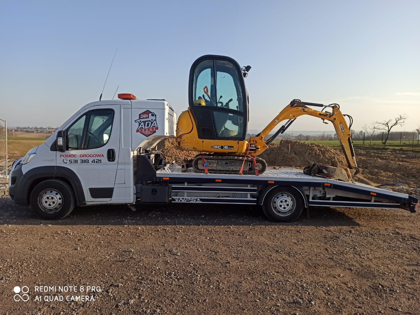 Biała laweta z żółtą mini koparką JCB 16CTS, zabezpieczoną czerwonymi pasami transportowymi, na tle brązowego pola i błękitnego nieba.