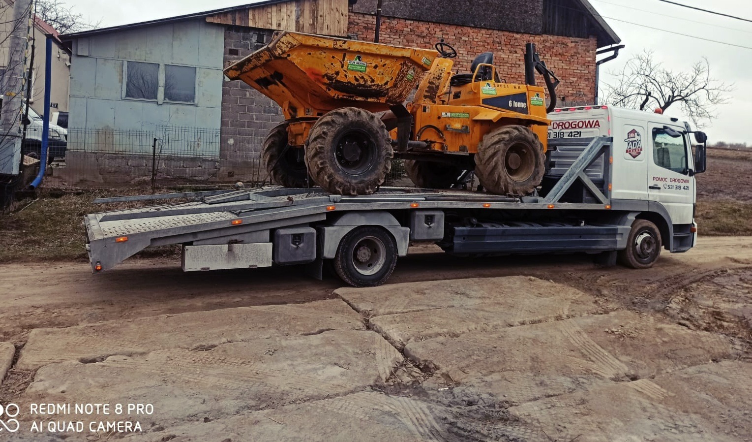 Żółta wywrotka budowlana 6-tonowa przewożona na lawecie pomocy drogowej z widocznym numerem telefonu i logo firmy na białej kabinie, w tle budynki gospodarcze.