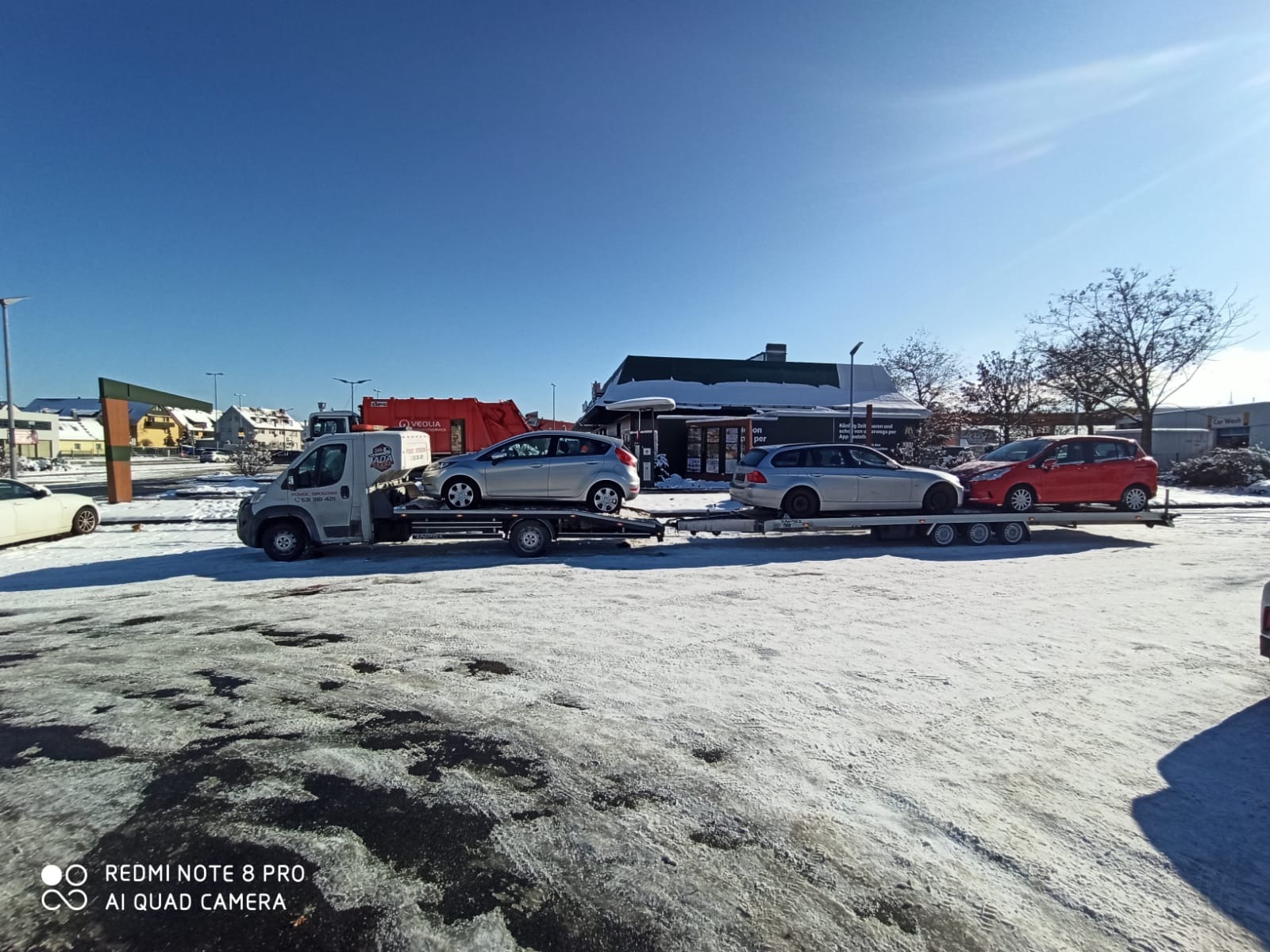Laweta z trzema samochodami różnych marek na parkingu pokrytym śniegiem w słoneczny dzień.