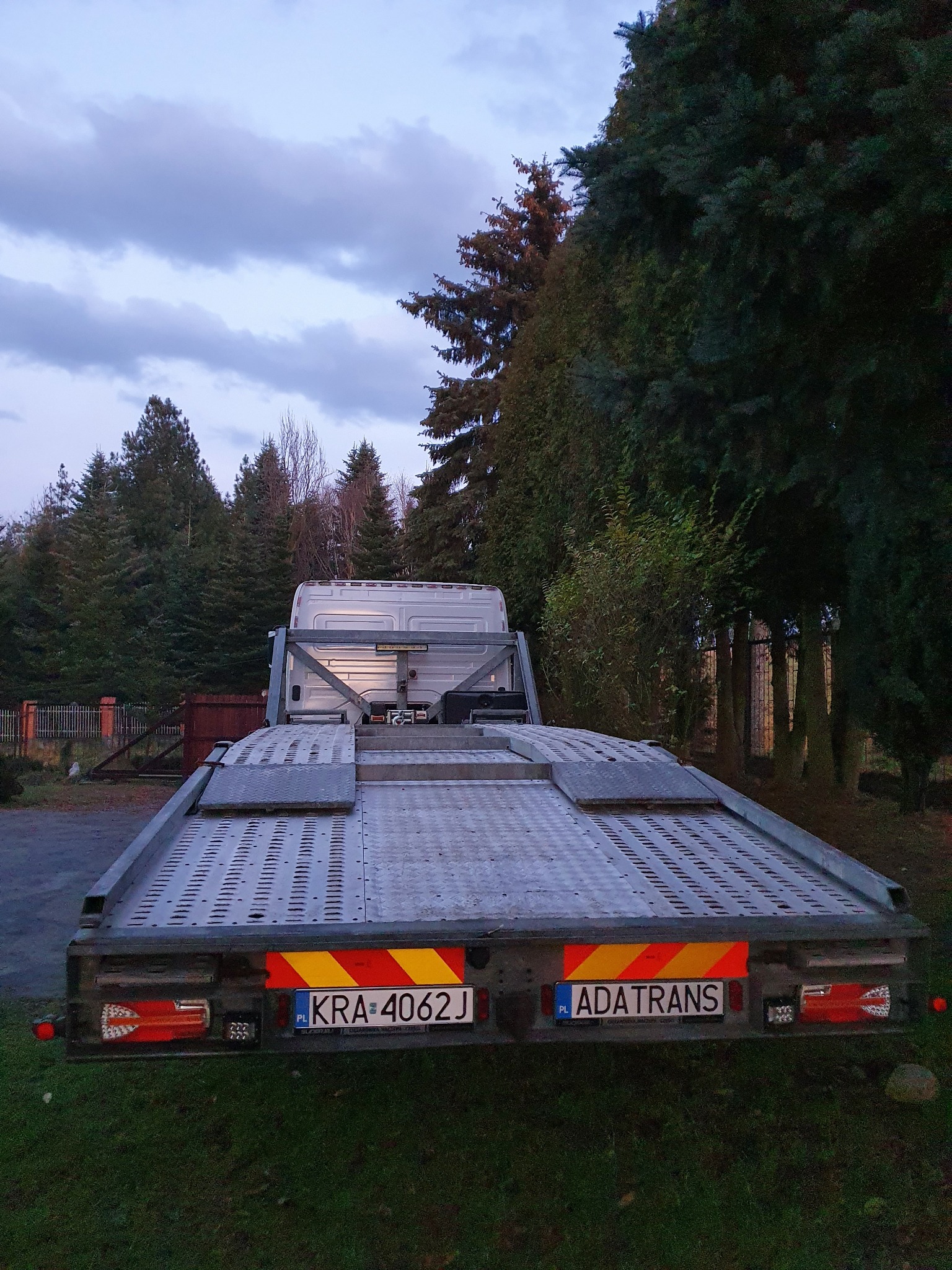 Pusta platforma lawety samochodowej z widocznymi tablicami rejestracyjnymi KRA-4062J i ADATRANS, zaparkowana na trawie w tle drzew i pochmurnego nieba o zmierzchu.