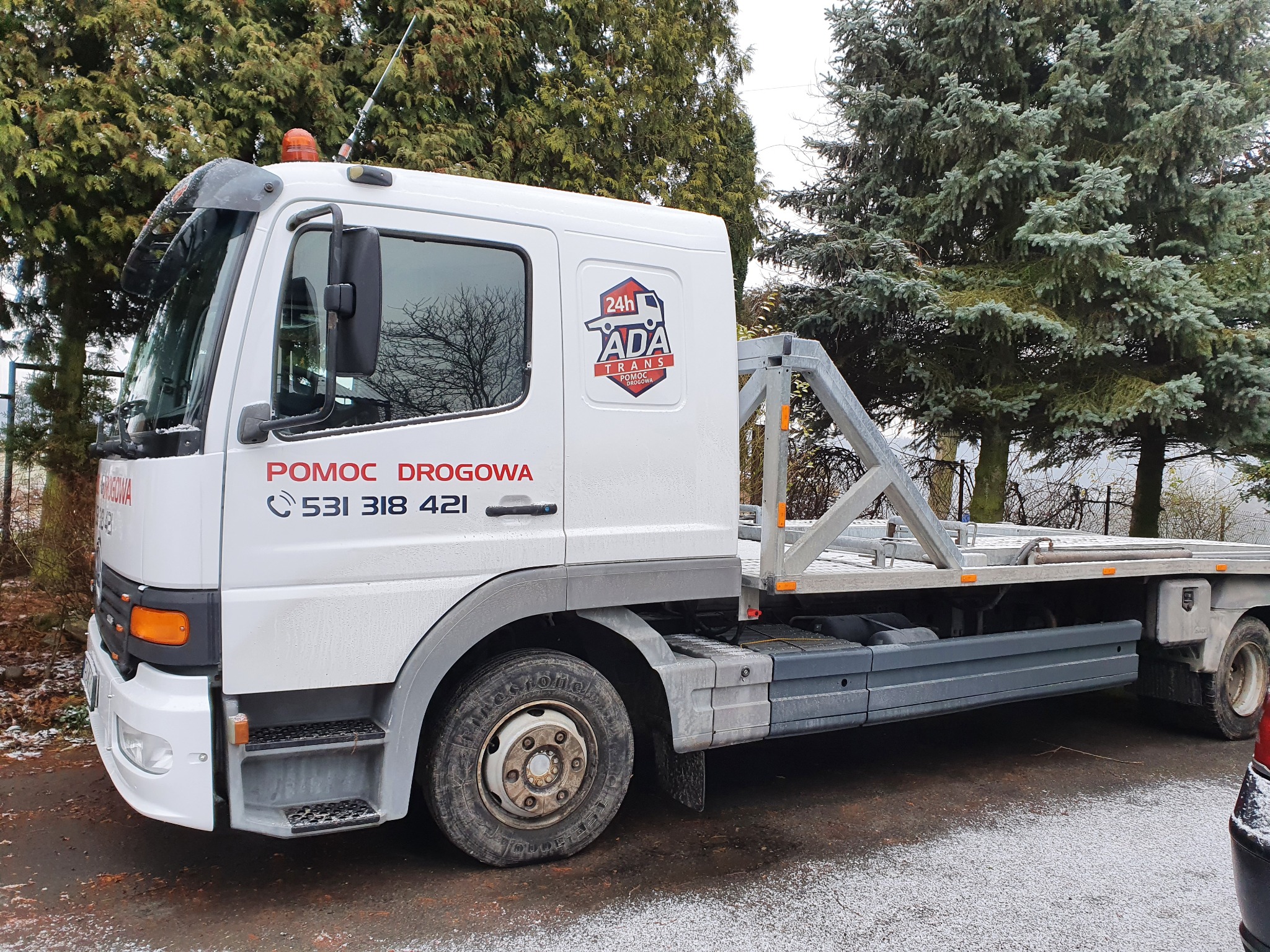 Biała laweta pomocy drogowej z numerem telefonu i logo firmy ADA Trans na tle zielonych drzew.