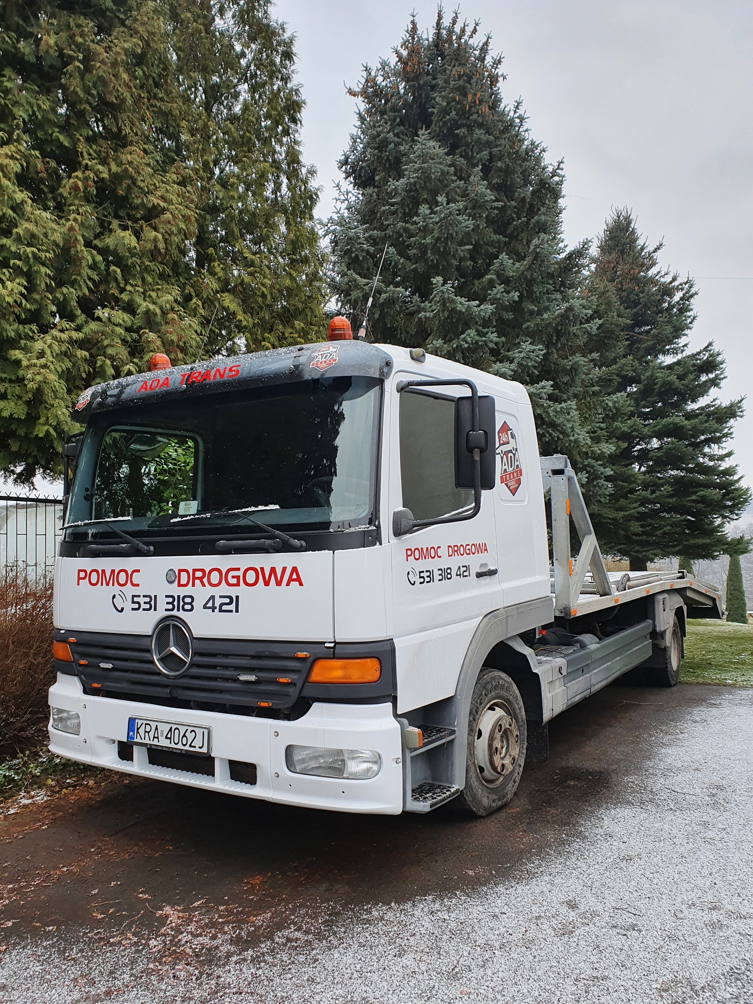 Biała laweta Mercedes-Benz z napisem 'Pomoc Drogowa' i numerem telefonu, zaparkowana na mokrym asfalcie przed zielonymi drzewami w pochmurny dzień.