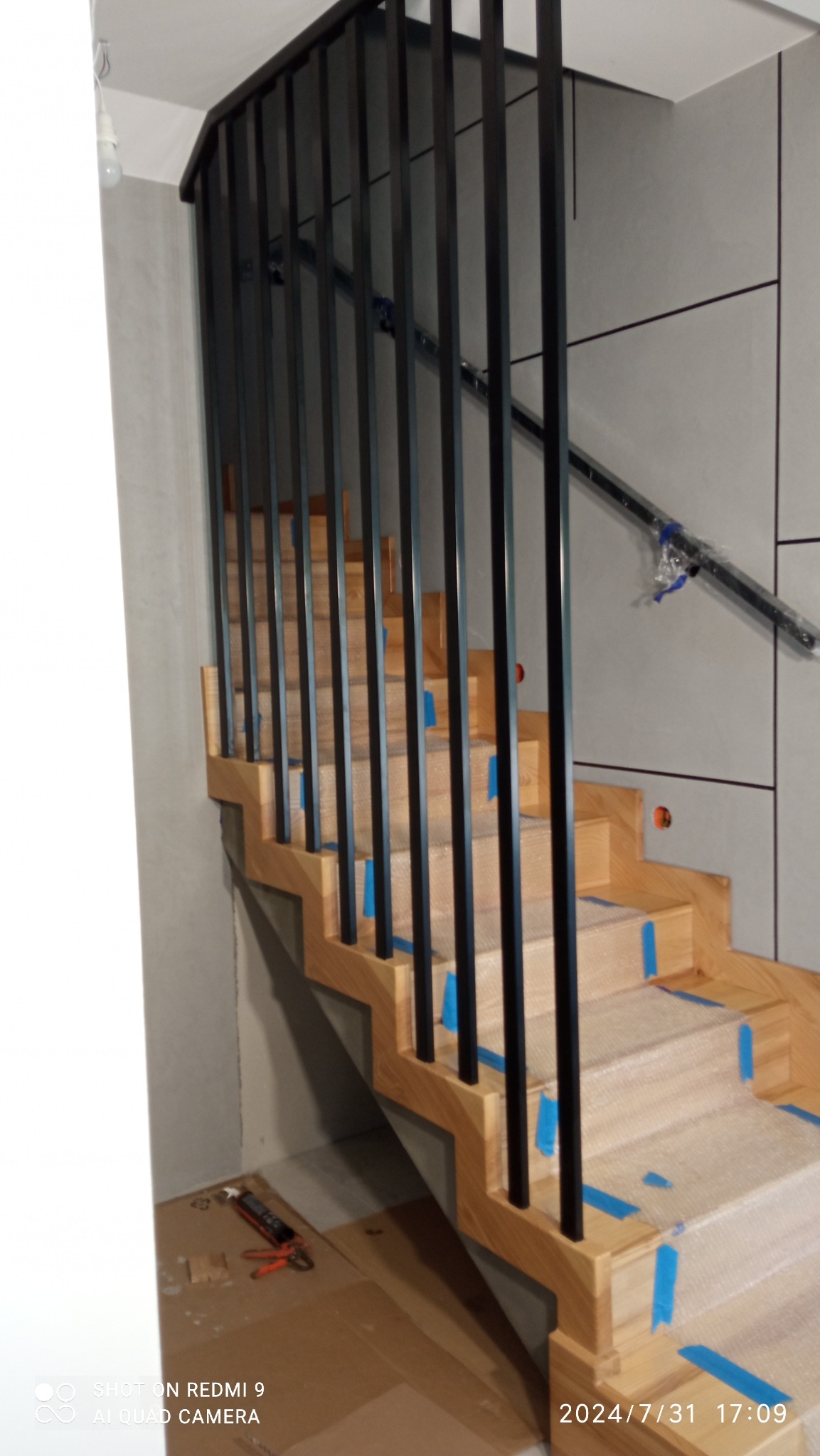 Montaż dębowych schodów z czarną, metalową balustradą w domu w Ładzicach, zabezpieczone folią i taśmą malarską na czas budowy.