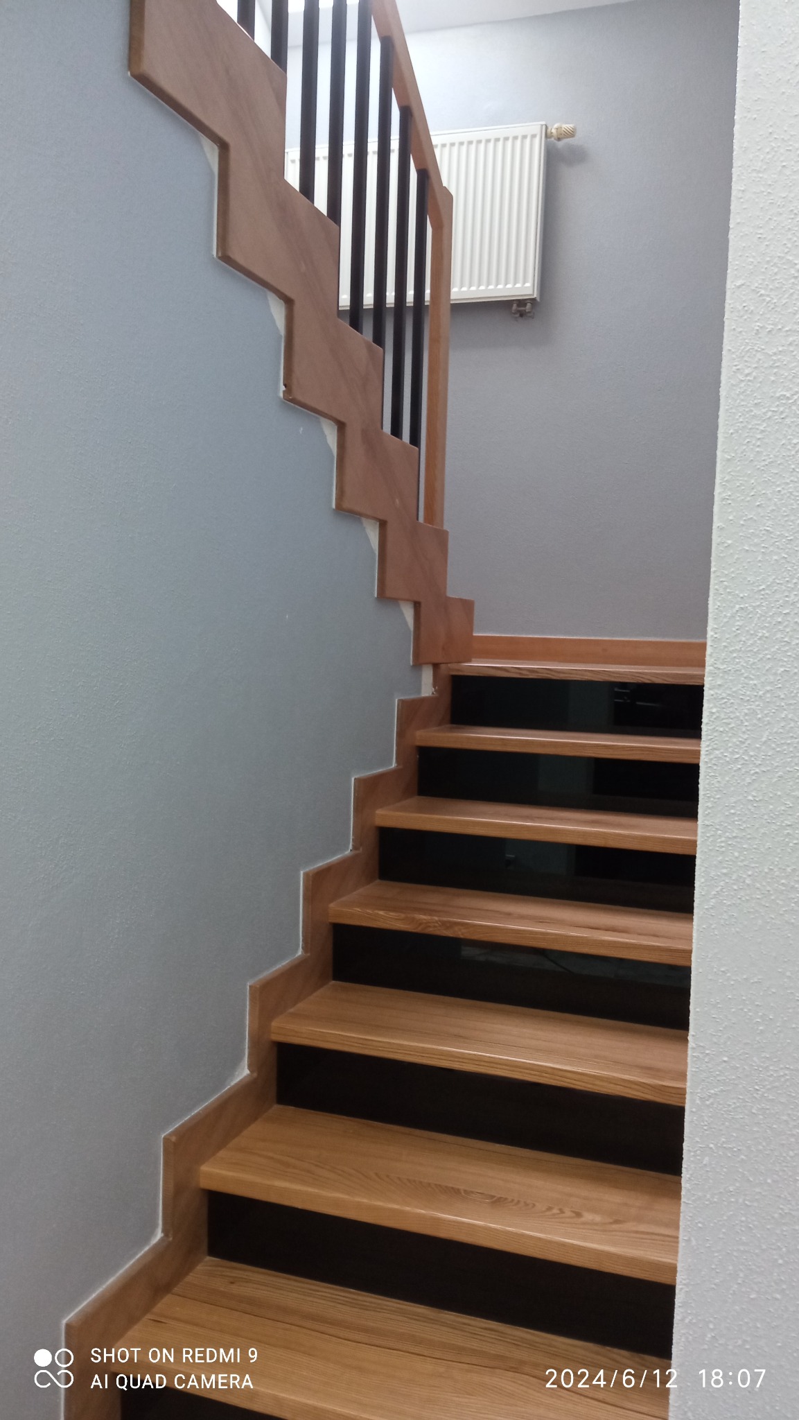 Drewniane schody z czarnymi balustradami na tle szarej ściany z grzejnikiem, widok z dołu.