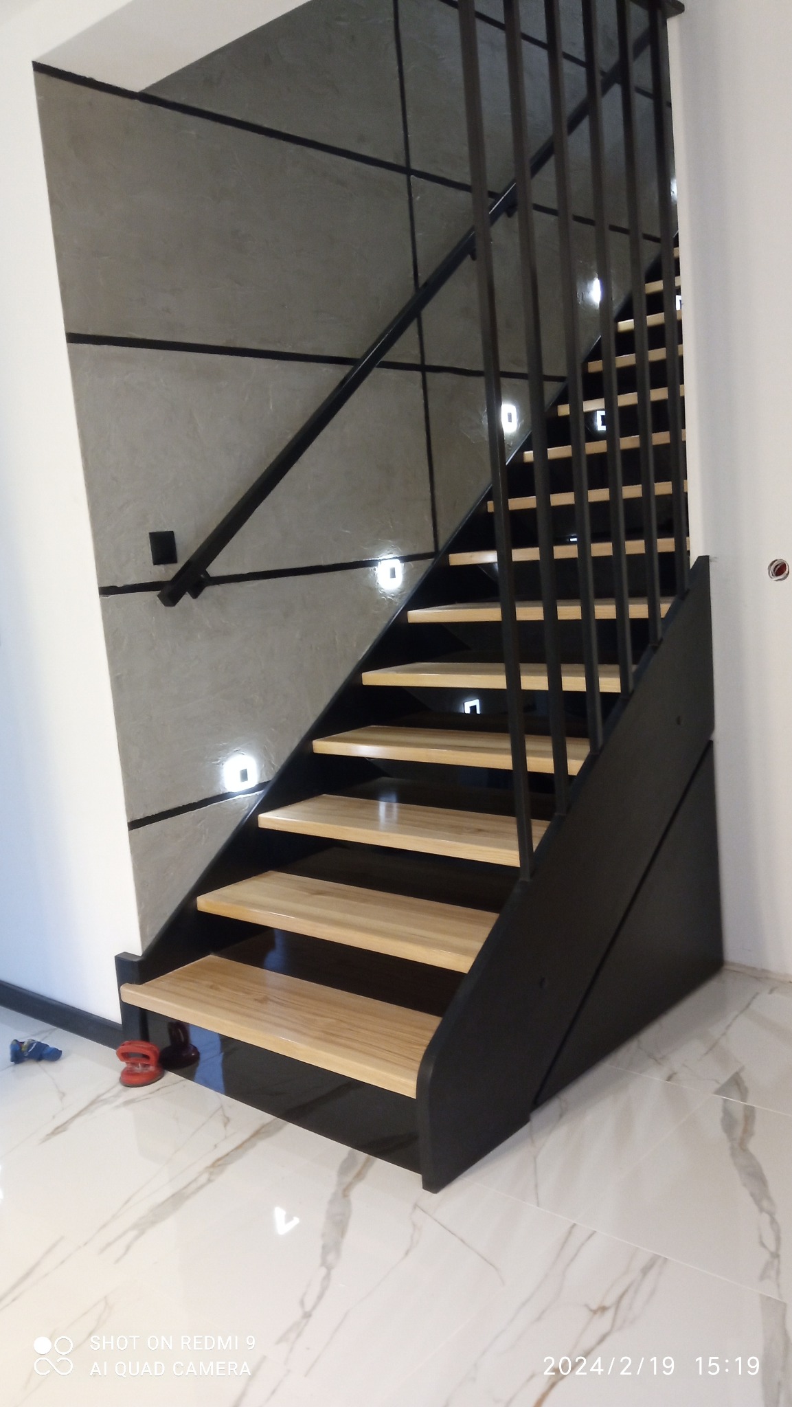 Nowoczesne schody z jasnego drewna i czarną metalową balustradą, oświetlone małymi kwadratowymi lampkami LED w ścianie, na marmurowej podłodze.