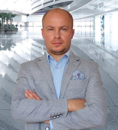 Portret mężczyzny w szarym blezerze i błękitnej koszuli, z rękami skrzyżowanymi na tle nowoczesnego, przeszklonego wnętrza biurowego.