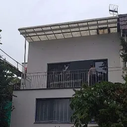 Dwupiętrowy dom murowany z płaskim dachem i zadaszeniem z poliwęglanu nad balkonem, widoczny fragment elewacji po remoncie z nowymi oknami i metalową balustradą, na dachu antena.