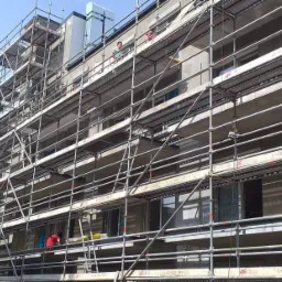 Budynek wielorodzinny w trakcie prac elewacyjnych, otoczony rusztowaniami, z widocznymi pracownikami na różnych poziomach.