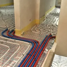 Rozłożona instalacja ogrzewania podłogowego z rurami w kolorach białym, niebieskim i czerwonym na folii izolacyjnej z siatką. Widoczne przejścia między pomieszczeniami.