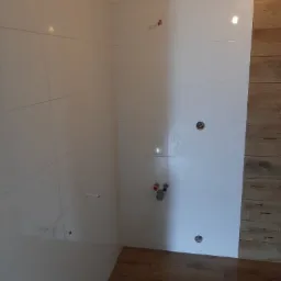 Ściana wyłożona białymi, błyszczącymi płytkami, z widocznymi otworami na armaturę i wystającymi rurkami. Na ścianie po prawej stronie widoczne płytki imitujące drewno. Podłoga również wyłożona...