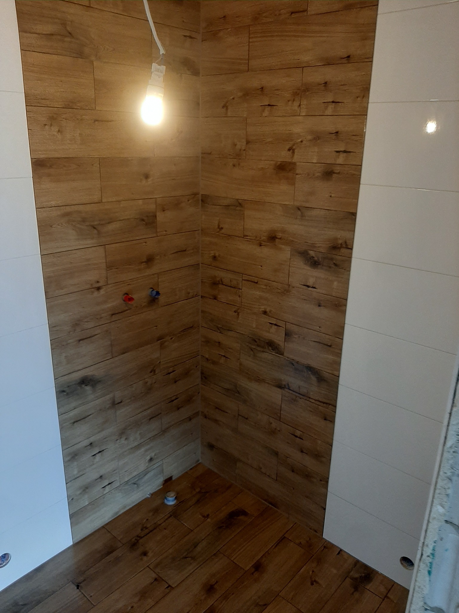 Wykończona kabina prysznicowa z drewnopodobnymi płytkami na ścianach i podłodze, kontrastująca z białymi płytkami po bokach, oświetlona wiszącą żarówką, widoczne przyłącza wodne.