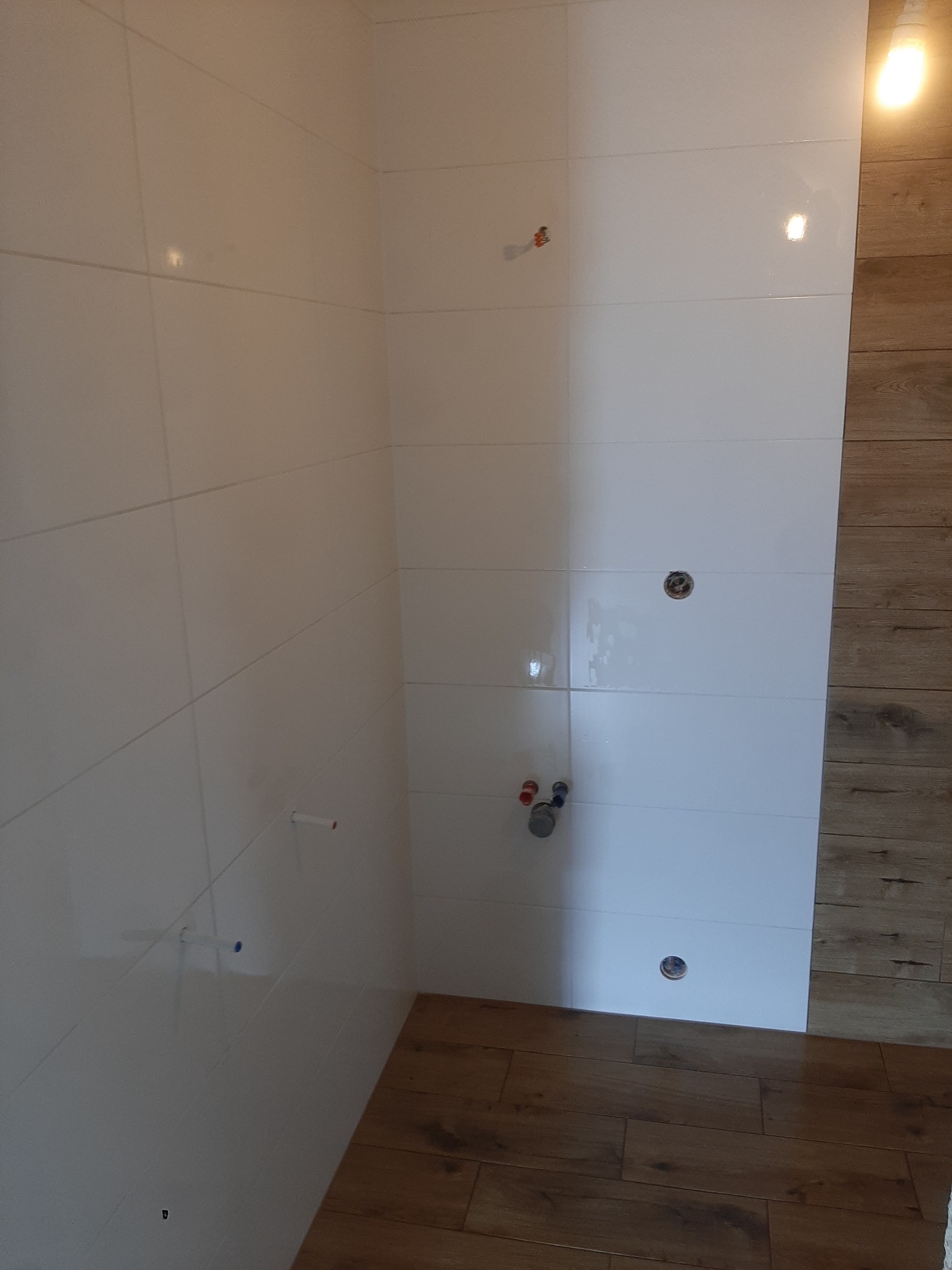 Ściana wyłożona białymi, błyszczącymi płytkami, z widocznymi otworami na armaturę i wystającymi rurkami. Na ścianie po prawej stronie widoczne płytki imitujące drewno. Podłoga również wyłożona...