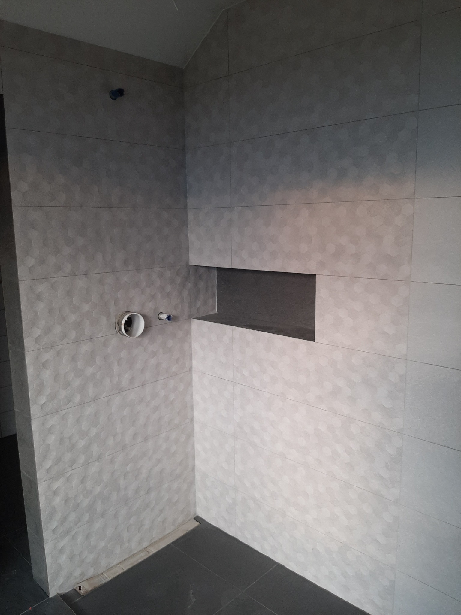 Szare płytki heksagonalne w trakcie montażu w łazience w Wiązowie, widoczna wnęka w ścianie i rury instalacyjne.