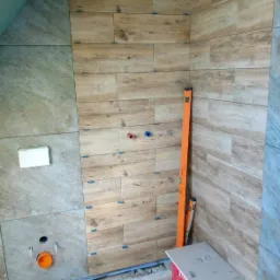 Montaż płytek imitujących drewno na ścianie w łazience, widoczne poziomice, krzyżyki dystansowe i przygotowane przyłącza wodne.