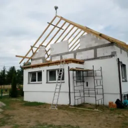 Budowa domu jednorodzinnego: ściany z betonu komórkowego, drewniana konstrukcja dachu, wieniec z kwiatami na szczycie, drabina przystawiona do ściany, rusztowanie przy elewacji.