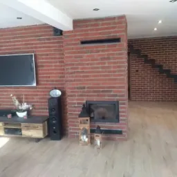 Salon z ceglaną ścianą, kominkiem i schodami z metalowymi stopniami, oświetlony punktowo, z widocznym sprzętem audio-wideo i drewnianą podłogą.