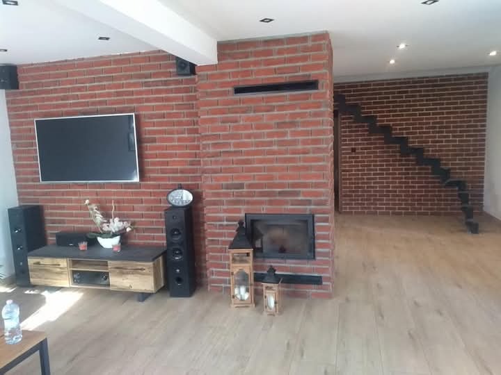 Salon z ceglaną ścianą, kominkiem i schodami z metalowymi stopniami, oświetlony punktowo, z widocznym sprzętem audio-wideo i drewnianą podłogą.