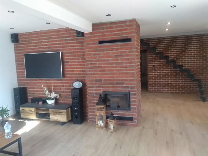 Salon z ceglaną ścianą, kominkiem, telewizorem i schodami, wykończony drewnianą podłogą, oświetlony punktowymi lampami sufitowymi.