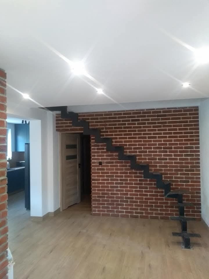 Nowoczesne, minimalistyczne schody metalowe bez poręczy, zamontowane na ścianie z czerwonej cegły w jasnym pomieszczeniu z drewnianą podłogą i białym sufitem z punktowym oświetleniem.