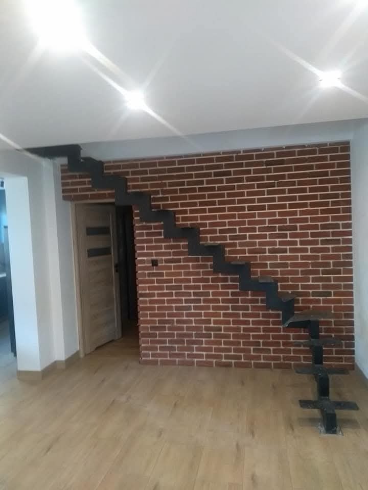 Minimalistyczne schody metalowe bez poręczy, zamontowane na tle ściany z czerwonej cegły w jasnym wnętrzu z panelami podłogowymi.