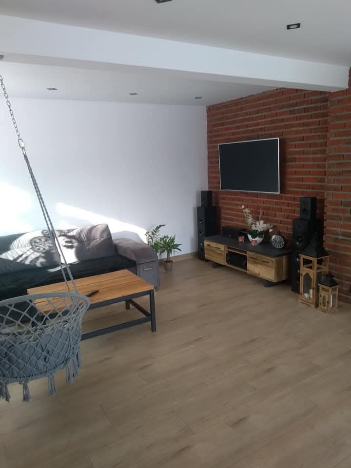 Nowoczesny salon z ceglaną ścianą, wiszącym fotelem, drewnianym stolikiem kawowym i podłogą z paneli w odcieniach beżu, oświetlony światłem dziennym wpadającym przez okno.