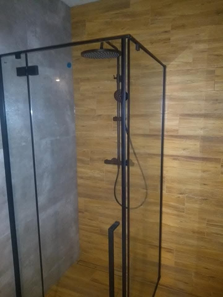 Nowoczesna kabina prysznicowa typu walk-in z czarną armaturą i szklanymi ściankami, wykończona płytkami imitującymi drewno i beton.