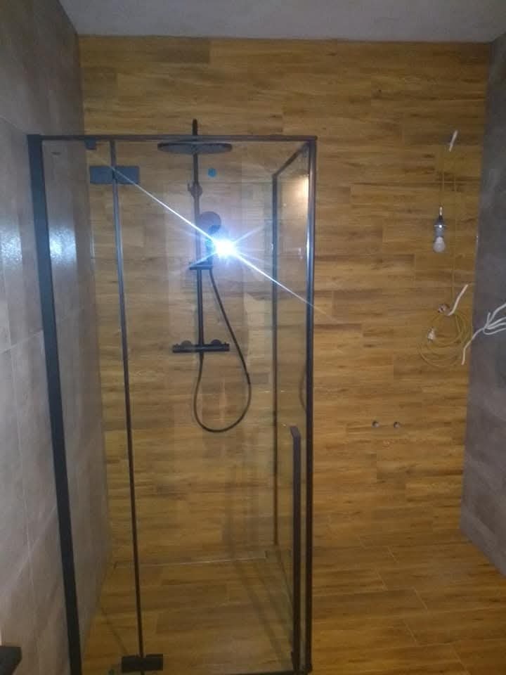 Nowoczesna kabina prysznicowa z czarnymi profilami, drewnopodobne płytki na ścianach i podłodze, widoczne niedokończone podłączenia elektryczne.