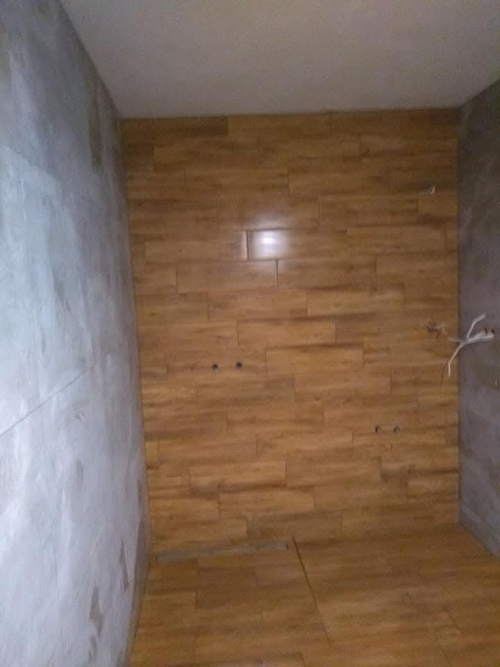 Ściana i podłoga w trakcie wykańczania płytkami imitującymi drewno, widoczne przewody elektryczne i surowa ściana obok.