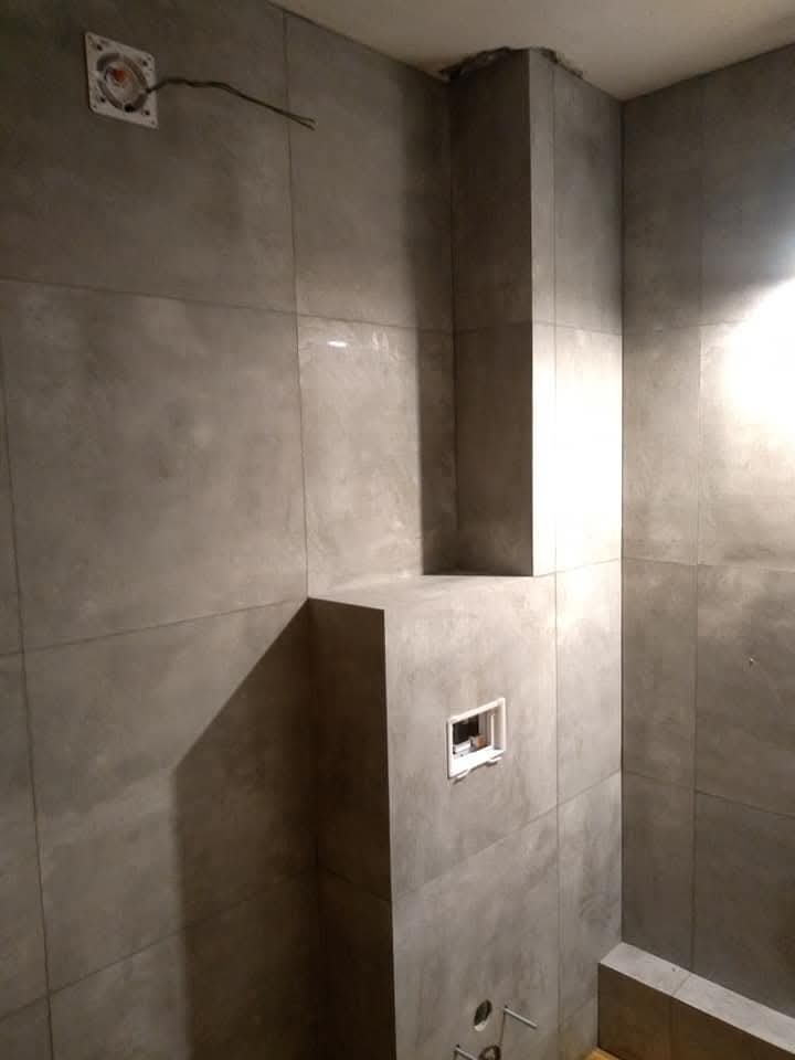 Świeżo wyłożona, szara, matowa glazura w łazience, z widocznymi wystającymi przewodami elektrycznymi i niedokończonymi otworami na armaturę, ukazująca etap prac wykończeniowych.