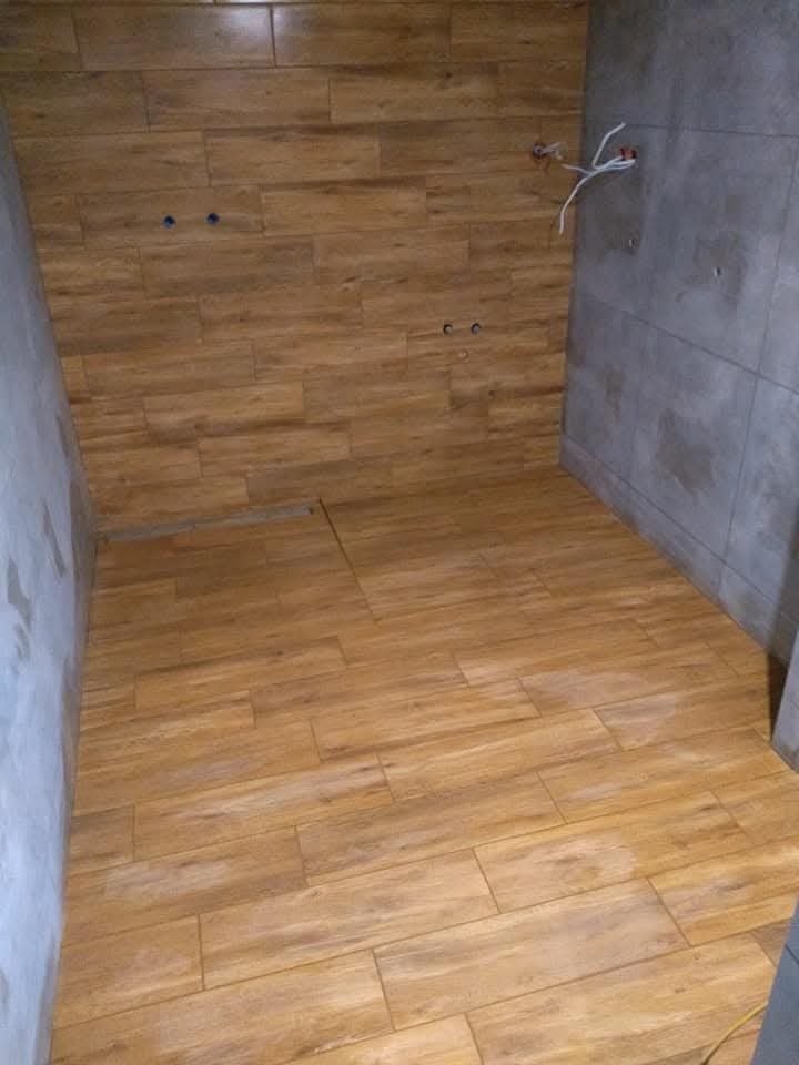 Wnętrze łazienki w trakcie wykańczania: drewnopodobne płytki na podłodze i ścianie, szare płytki na fragmencie ściany, wystające kable elektryczne, widoczne otwory na baterię.