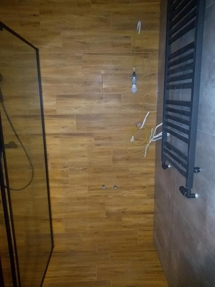 Wnętrze łazienki w trakcie remontu, widoczne płytki imitujące drewno na ścianach i podłodze, czarna kabina prysznicowa z lewej strony, grzejnik drabinkowy na ścianie z prawej, wisząca żarówka...