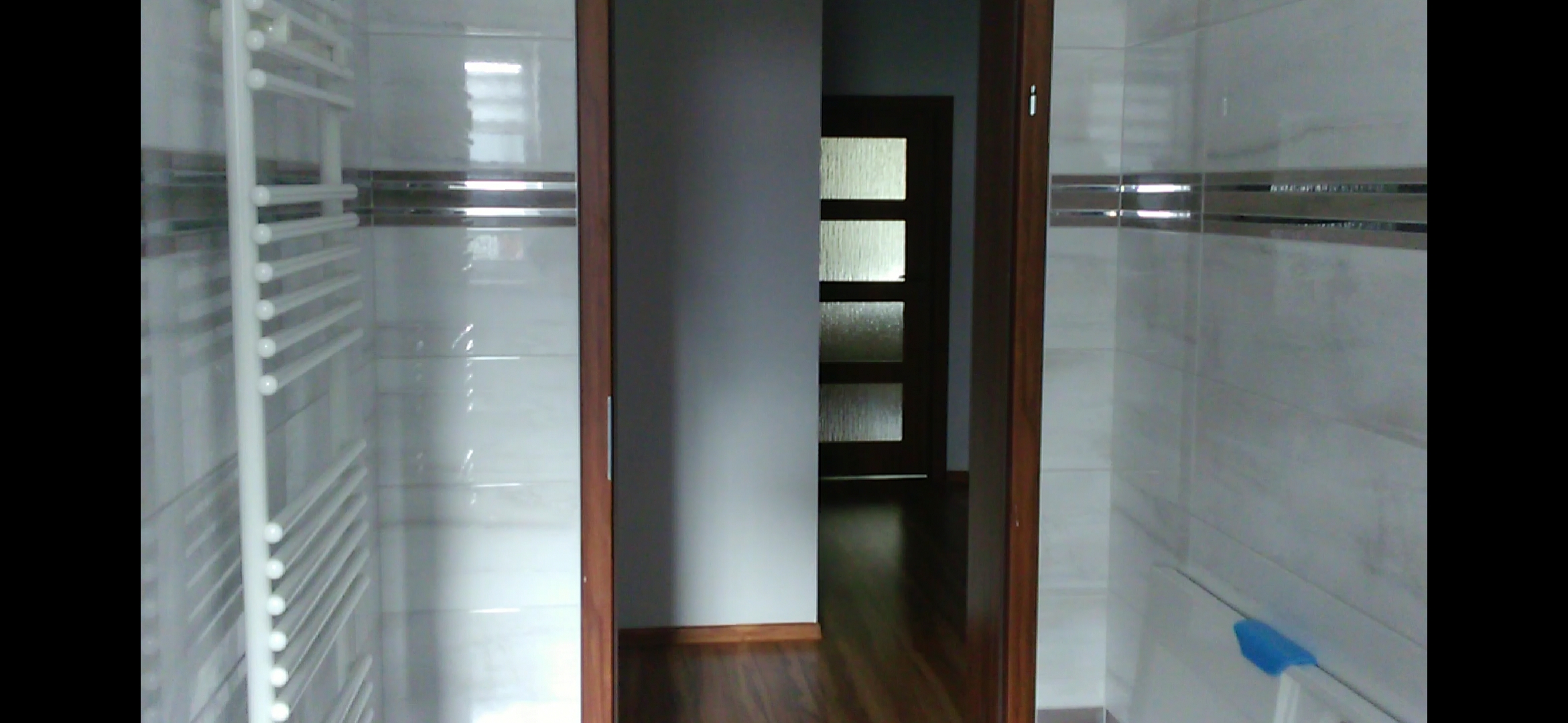 Wnętrze łazienki z jasnymi płytkami z metalicznymi paskami, grzejnik drabinkowy po lewej stronie i widok na pokój przez otwarte drewniane drzwi.