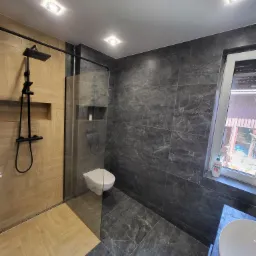 Nowoczesna łazienka z kabiną prysznicową typu walk-in, wykończona płytkami w odcieniach szarości i beżu, z czarną armaturą i odpływem liniowym w podłodze.