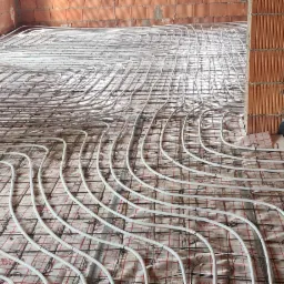 Instalacja ogrzewania podłogowego z rur PEX na siatce w surowym wnętrzu, widoczne czerwone rury doprowadzające i ściany z cegły dziurawki.