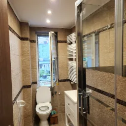 Wyremontowana łazienka z beżowymi płytkami, otwartym oknem z widokiem na budynek i grzejnikiem drabinkowym, kabina prysznicowa z widocznym odbiciem.