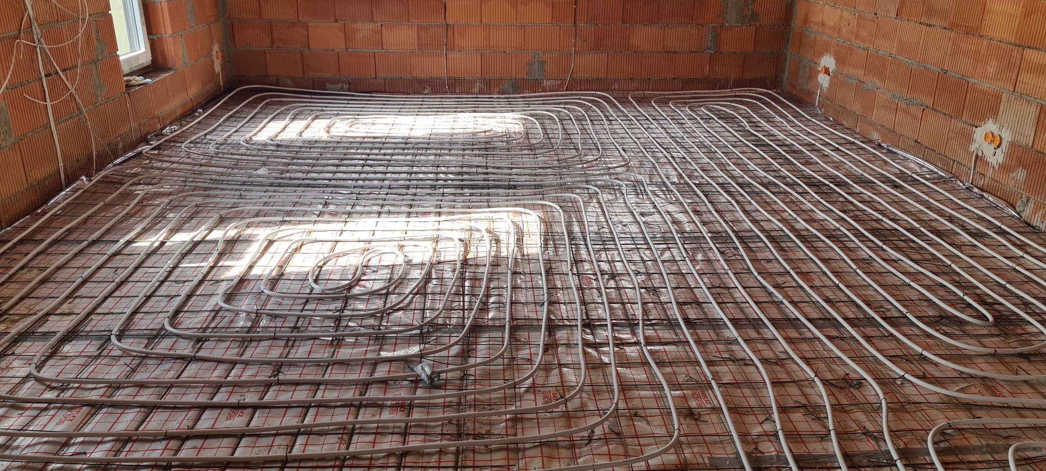 Rozłożone rury ogrzewania podłogowego na izolacji w surowym wnętrzu z ceglanymi ścianami i widocznymi puszkami elektrycznymi.
