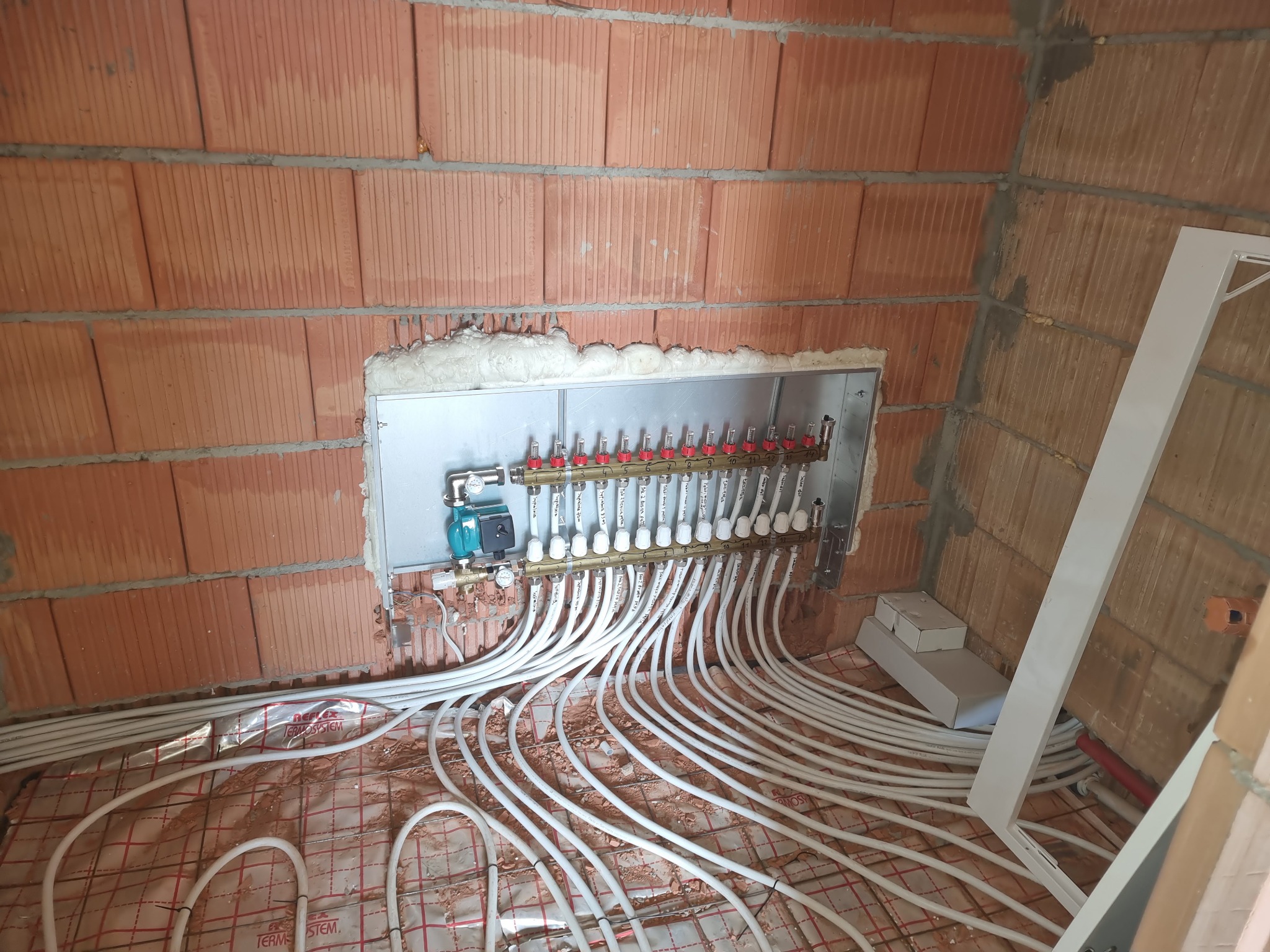 Rozdzielacz ogrzewania podłogowego z licznymi pętlami rur na siatce, zamontowany na ścianie z cegły w surowym stanie.