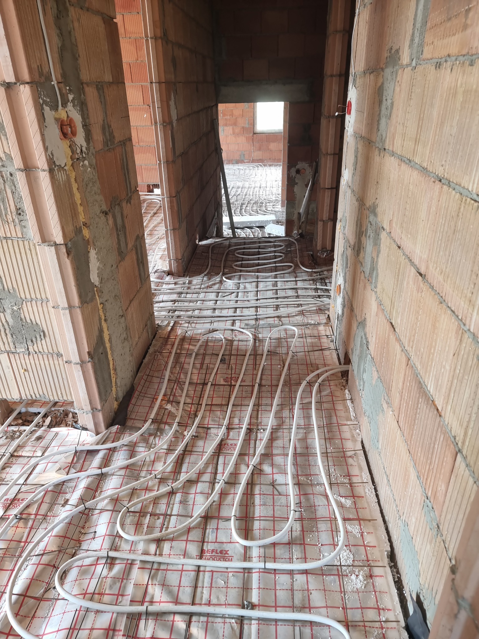 Wnętrze w trakcie budowy z widoczną instalacją ogrzewania podłogowego na folii z siatką, rury ułożone w pętle między surowymi ścianami z cegły.