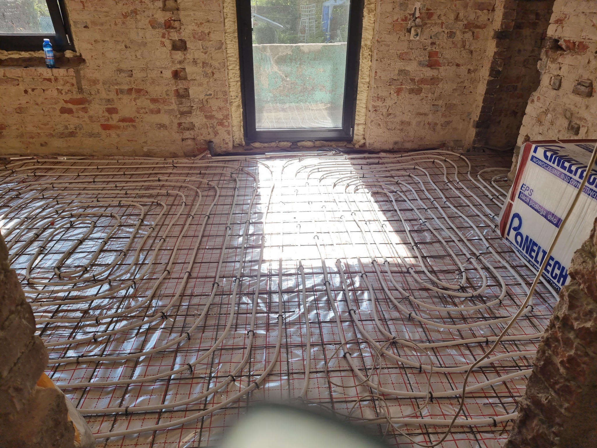 Białe rury ogrzewania podłogowego ułożone na folii z siatką w surowym pomieszczeniu z ceglanymi ścianami i oknem, widoczny fragment styropianu. W tle butelka płynu.