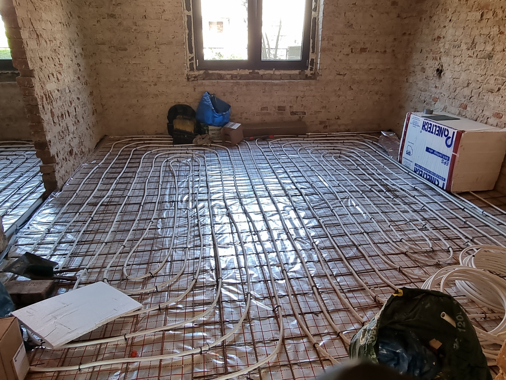 Montaż ogrzewania podłogowego: rury grzewcze ułożone na metalowej siatce na folii izolacyjnej w surowym pomieszczeniu z ceglanymi ścianami, widoczne materiały budowlane i narzędzia.