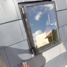 Montaż okna dachowego na szarym dachu z blachy trapezowej. Widoczny fragment narzędzia, czerwony przewód i odbicie nieba w szybie.