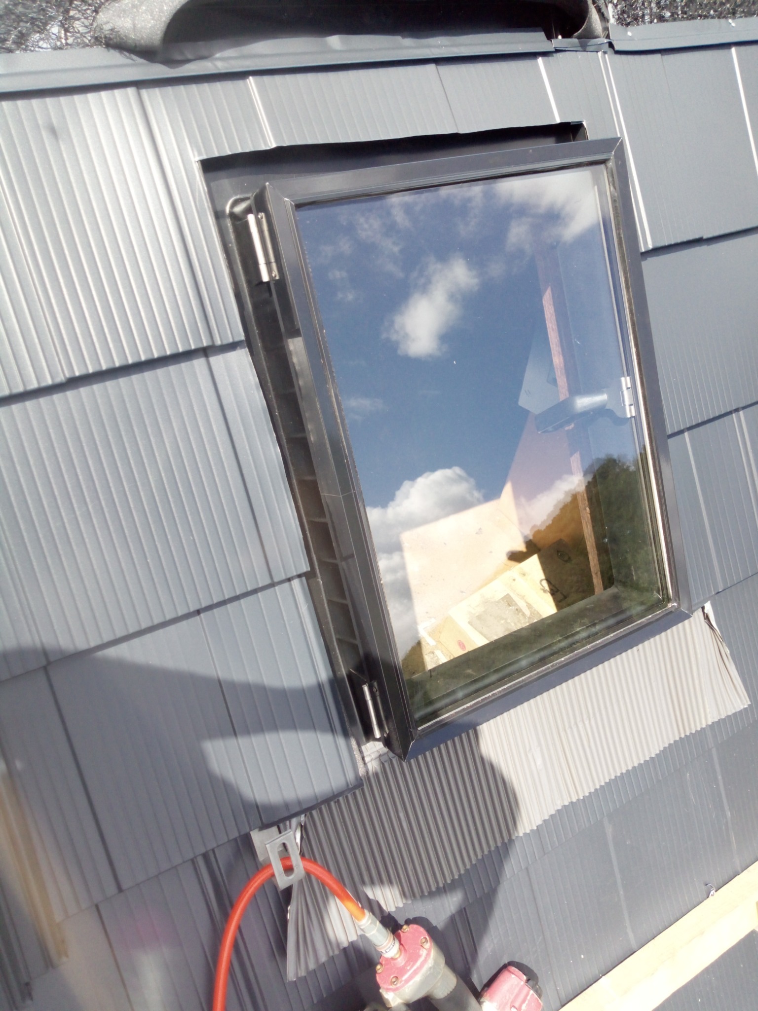 Montaż okna dachowego na szarym dachu z blachy trapezowej. Widoczny fragment narzędzia, czerwony przewód i odbicie nieba w szybie.