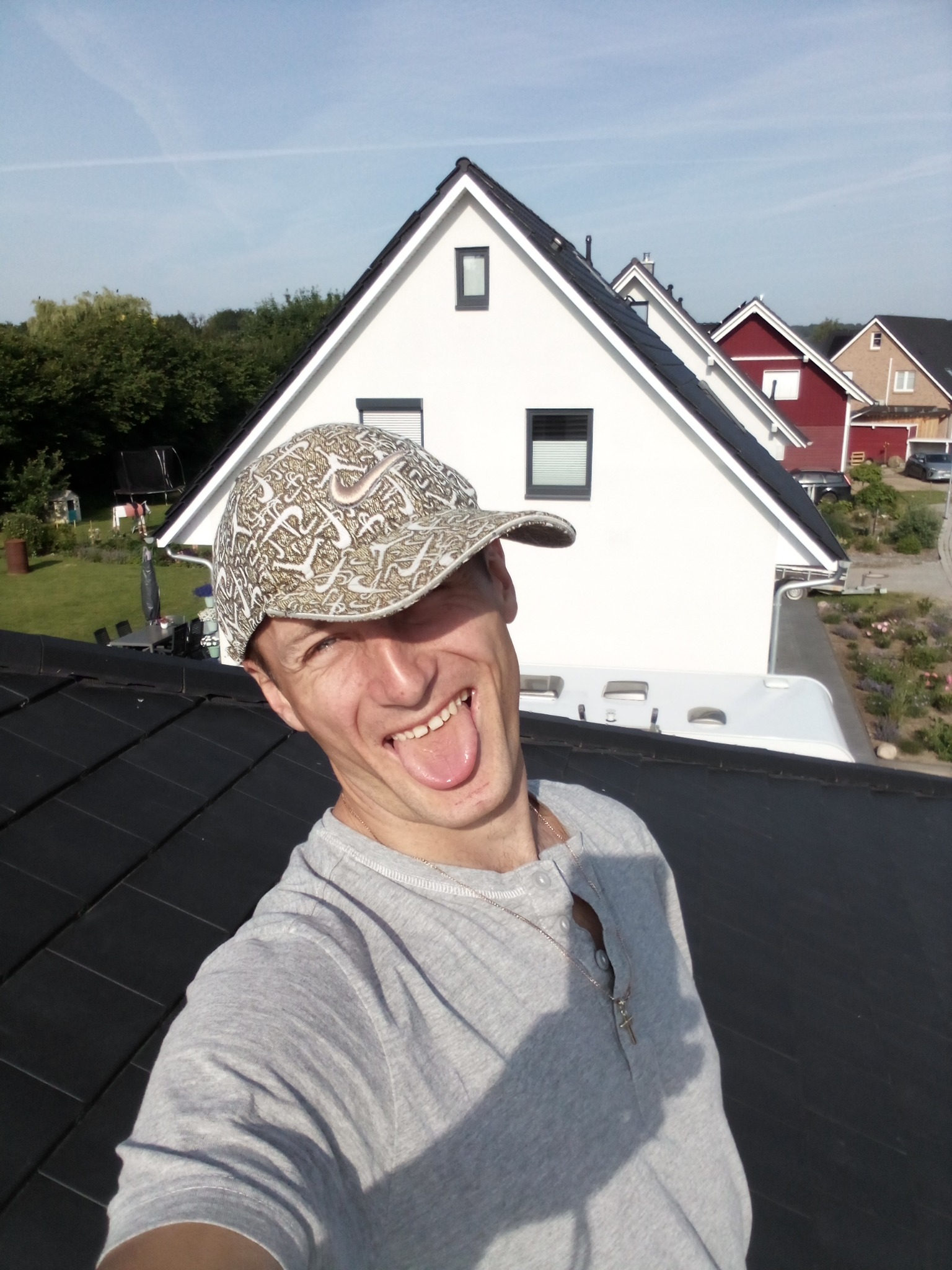 Wesoły mężczyzna w czapce robi selfie na tle czarnego dachu, pokazując język. W tle domy i błękitne niebo.