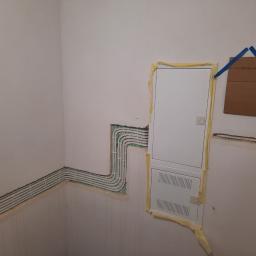 Lew instal - Ściana w trakcie remontu z zamontowanymi białymi korytkami kablowymi i białą skrzynką elektryczną zabezpieczoną taśmą malarską, na ścianie kartka z napisem 'K-KLIMATYZACJA' przyklejona niebieską...