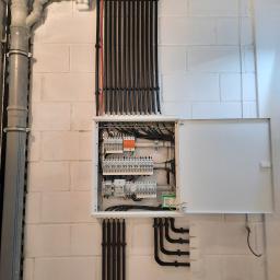 Lew instal - Otwarta skrzynka rozdzielcza z widocznymi bezpiecznikami i przewodami, zamontowana na pomalowanej na biało ścianie z bloczków betonowych. Rury instalacyjne w kolorze czarnym prowadzone pionowo...