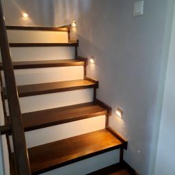 Lew instal - Drewniane schody z białymi podstopnicami, oświetlone małymi, kwadratowymi lampkami ściennymi, zamontowanymi przy każdym stopniu. Widoczny włącznik światła na szarej ścianie.